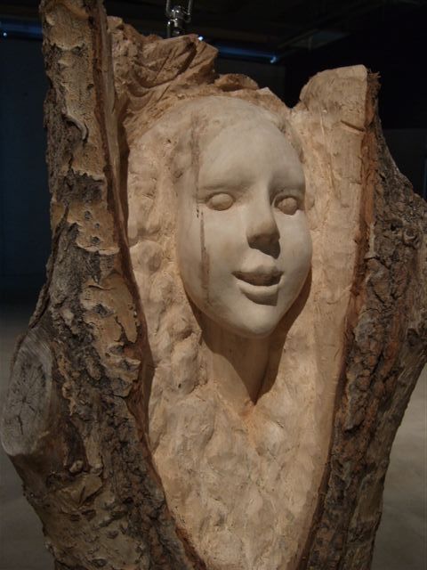 Close-up of a carved female face emerging from a tree trunk sculpture by Sanna Karlsson-Sutisna, from the Iroko exhibition at EMMA – Espoo Museum of Modern Art, 2010.