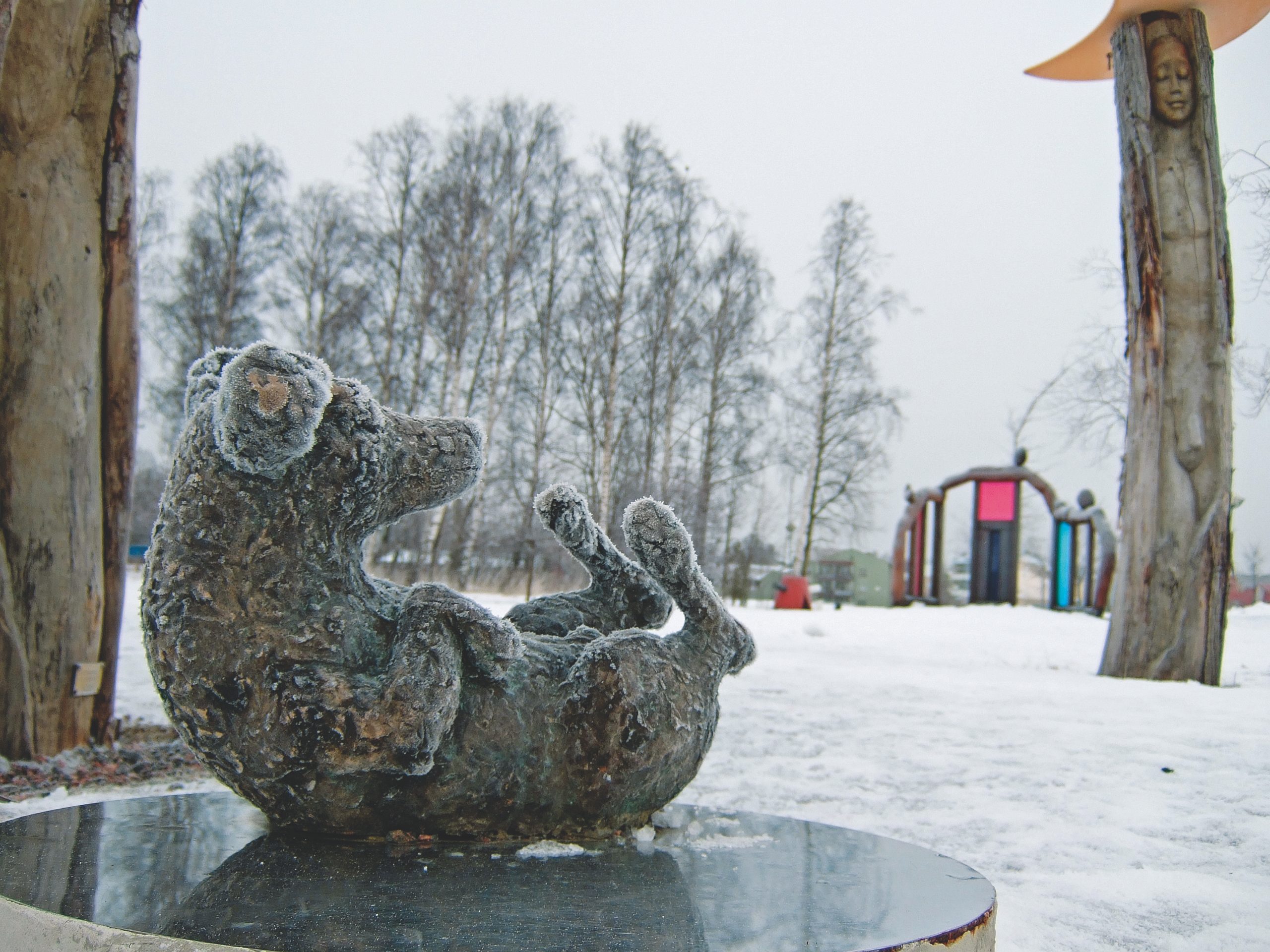 Public Artwork - Flower of Life - A frost-covered bronze armadillo sculpture on a pedestal with wooden sculptures in a snowy park. - Art park, Pikku-Huopalahti, Helsinki, 2005.
