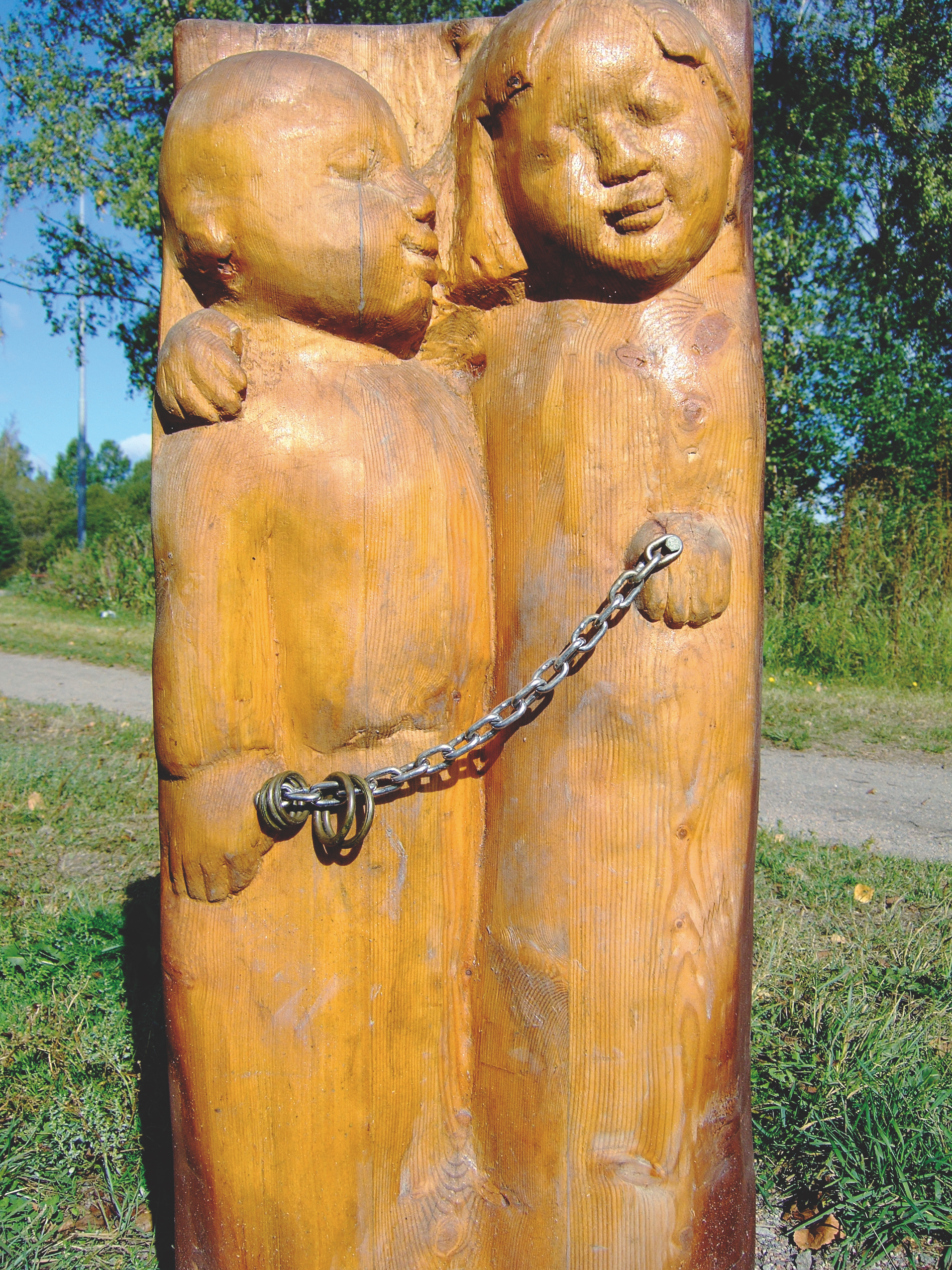 Public Artwork - Flower of Life - Carved wooden figures of two children linked by a metal chain, standing together in a grassy park. - Art park, Pikku-Huopalahti, Helsinki, 2005.