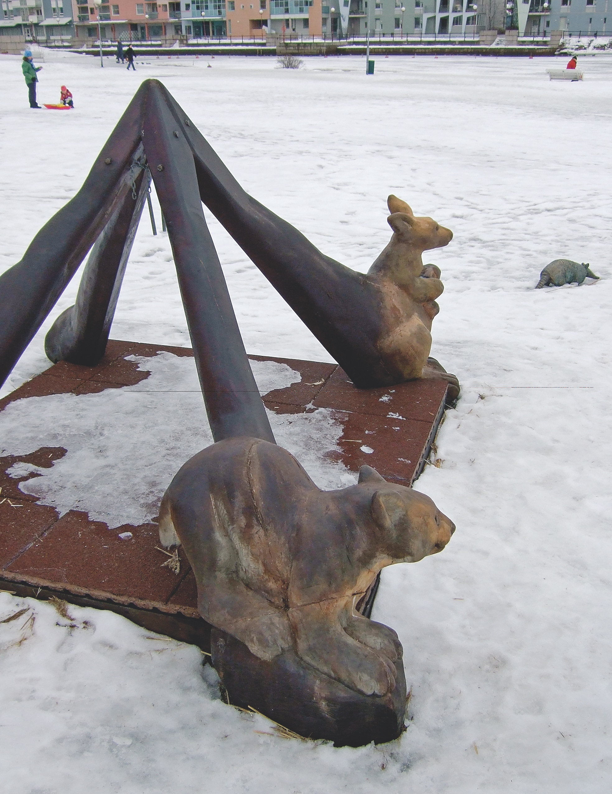 Public Artwork - Flower of Life - Wooden animal sculptures surrounding a play structure in a snowy park, with a bronze armadillo sculpture nearby. - Art park, Pikku-Huopalahti, Helsinki, 2005.