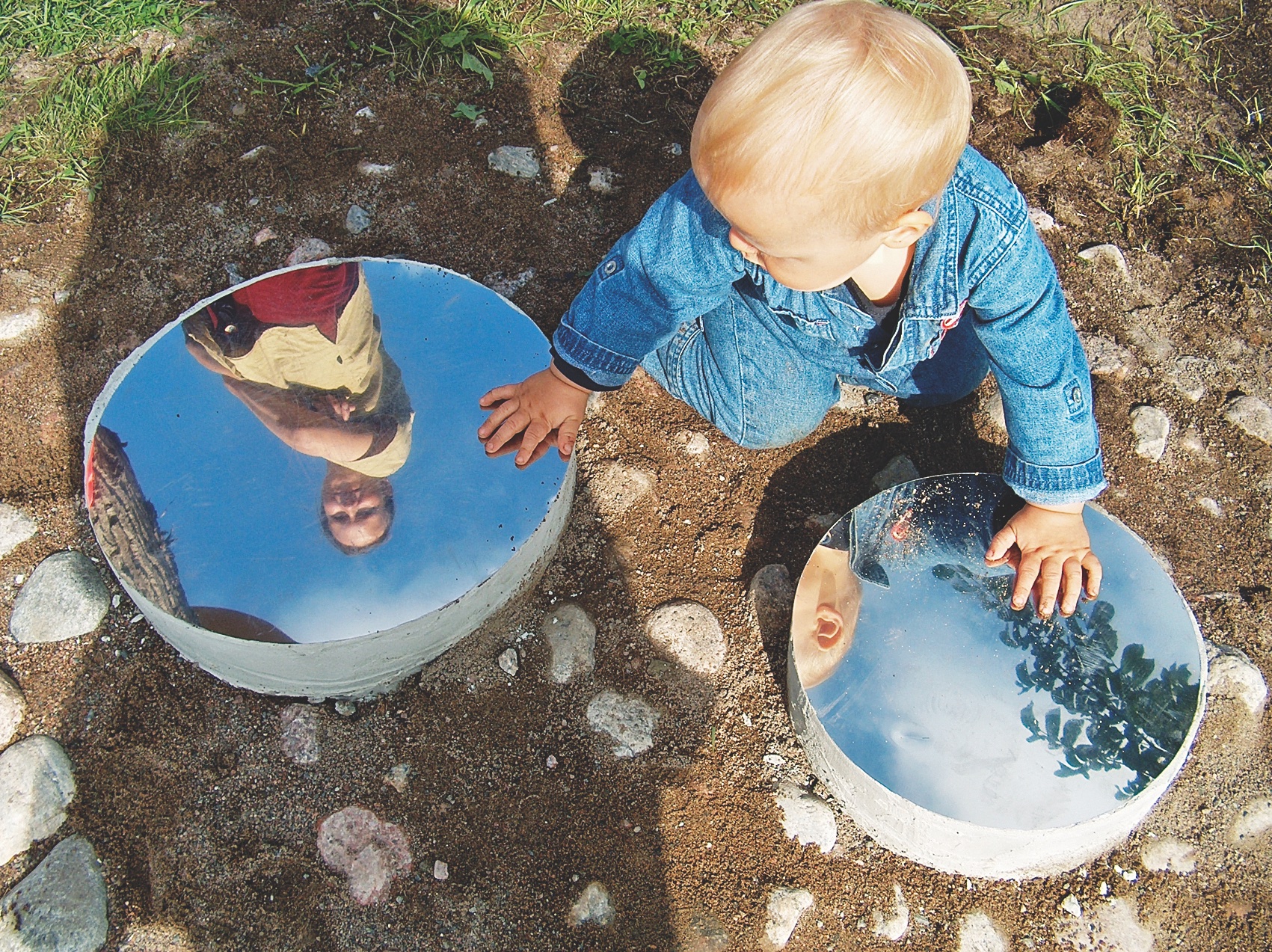 Public Artwork - Flower of Life - A child exploring two circular mirrored sculptures set into the ground in a grassy park. - Art park, Pikku-Huopalahti, Helsinki, 2005.