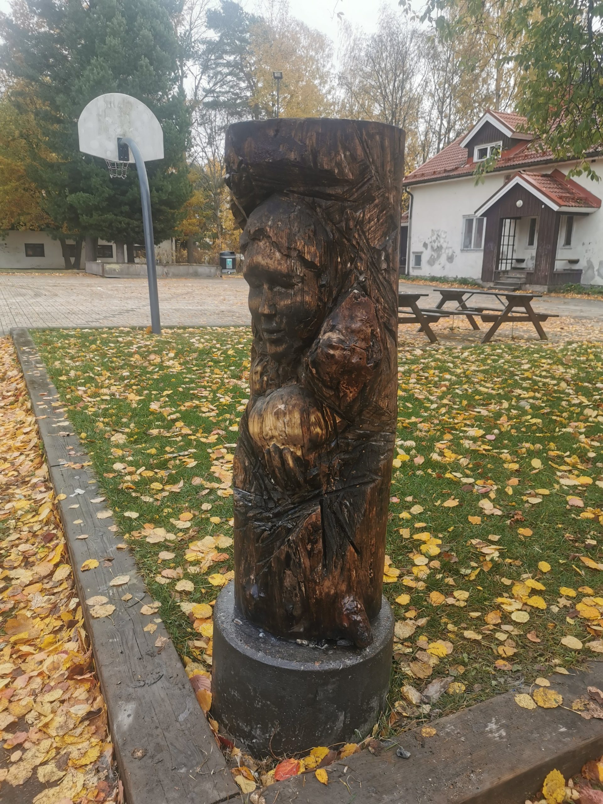 Public Artwork - A Maid and Child - Piikapuisto, Helsinki 2022. A carved wooden sculpture of a young girl holding an apple, carved into a tree trunk and mounted on a stone pedestal in an autumn park.