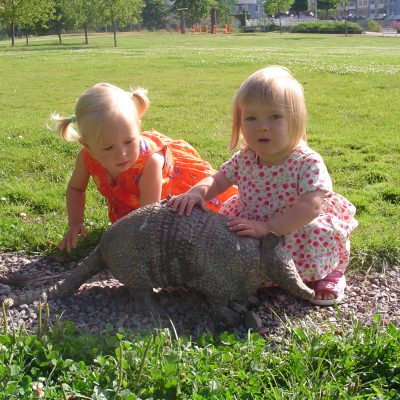 Public Artwork - Flower of Life - Two young children gently touching and exploring a bronze armadillo sculpture in a grassy park. - Art park, Pikku-Huopalahti, Helsinki, 2005.
